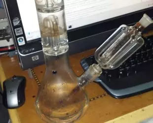 A glass water pipe standing on a desk in front of a laptop and keyboard.
