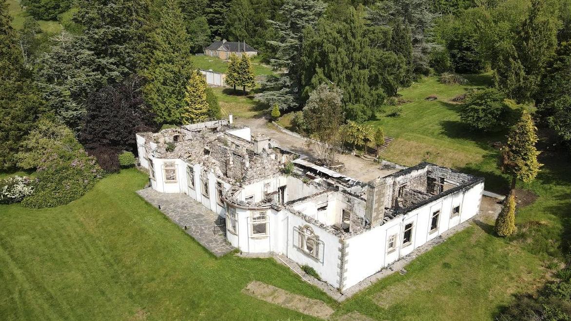 Aerial view of a fire-gutted country house, roof collapsed, still standing in a mown lawn ringed by trees.