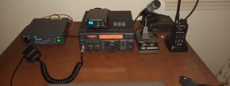 A ham radio bench: HF transceiver with lit frequency display, power supply, desk microphone and a handheld on its charger.